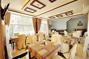 A dining area with tables set for four, chairs covered in white, and a large window with curtains. - Golden Classic Hotel Bagan (Nyaung-U)