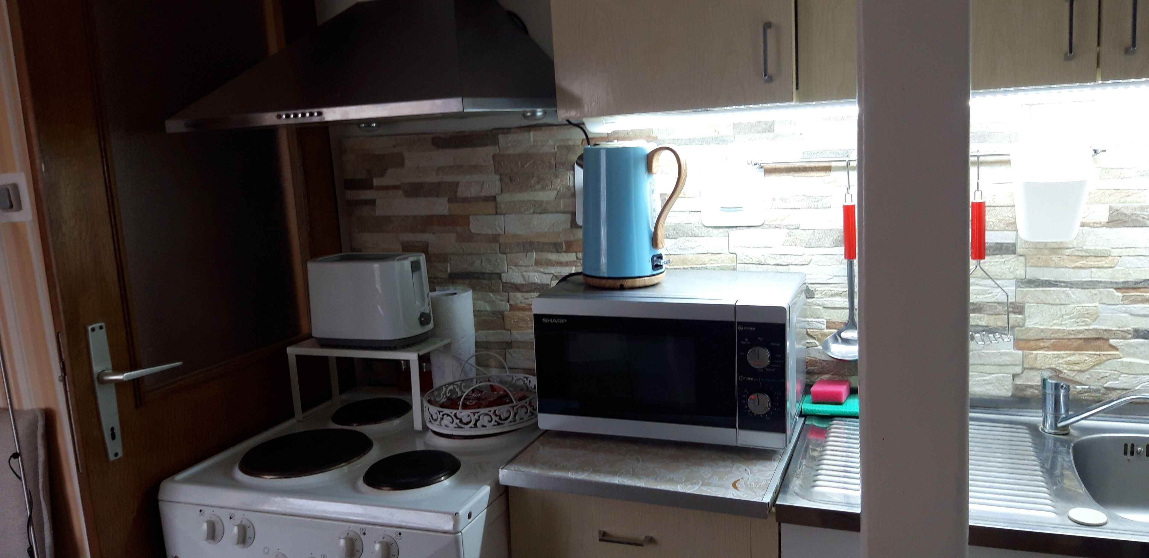 Een kleine keuken met een kookplaat, magnetron, waterkoker en een achterwand van steen.