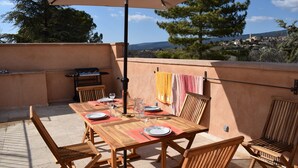 Una terrassa al terrat amb una taula de fusta preparada per dinar, una barbacoa i vistes del paisatge circumdant.