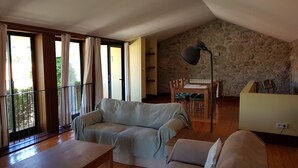 Ein Wohnzimmer mit Sofa, Esstisch und Lampe. Eine Steinwand und ein Balkon mit Vorhängen sind zu sehen.
