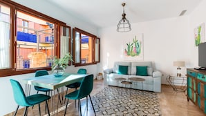 Un soggiorno moderno con zona pranzo, divano e televisore. È presente una grande finestra con vista su un edificio blu e un balcone.