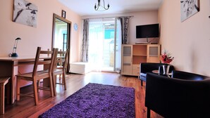 Uma sala de estar com uma mesa de jantar de madeira, cadeiras, um sofá preto, um tapete roxo, uma TV sobre um suporte e uma porta que dá para o exterior.