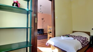 Quarto com cama, uma cadeira de madeira, prateleira com objeto decorativo e porta que dá para outro cômodo.