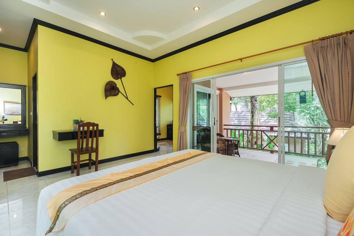 A bedroom with a large bed, a desk, and a chair. There is a balcony with a view of greenery.