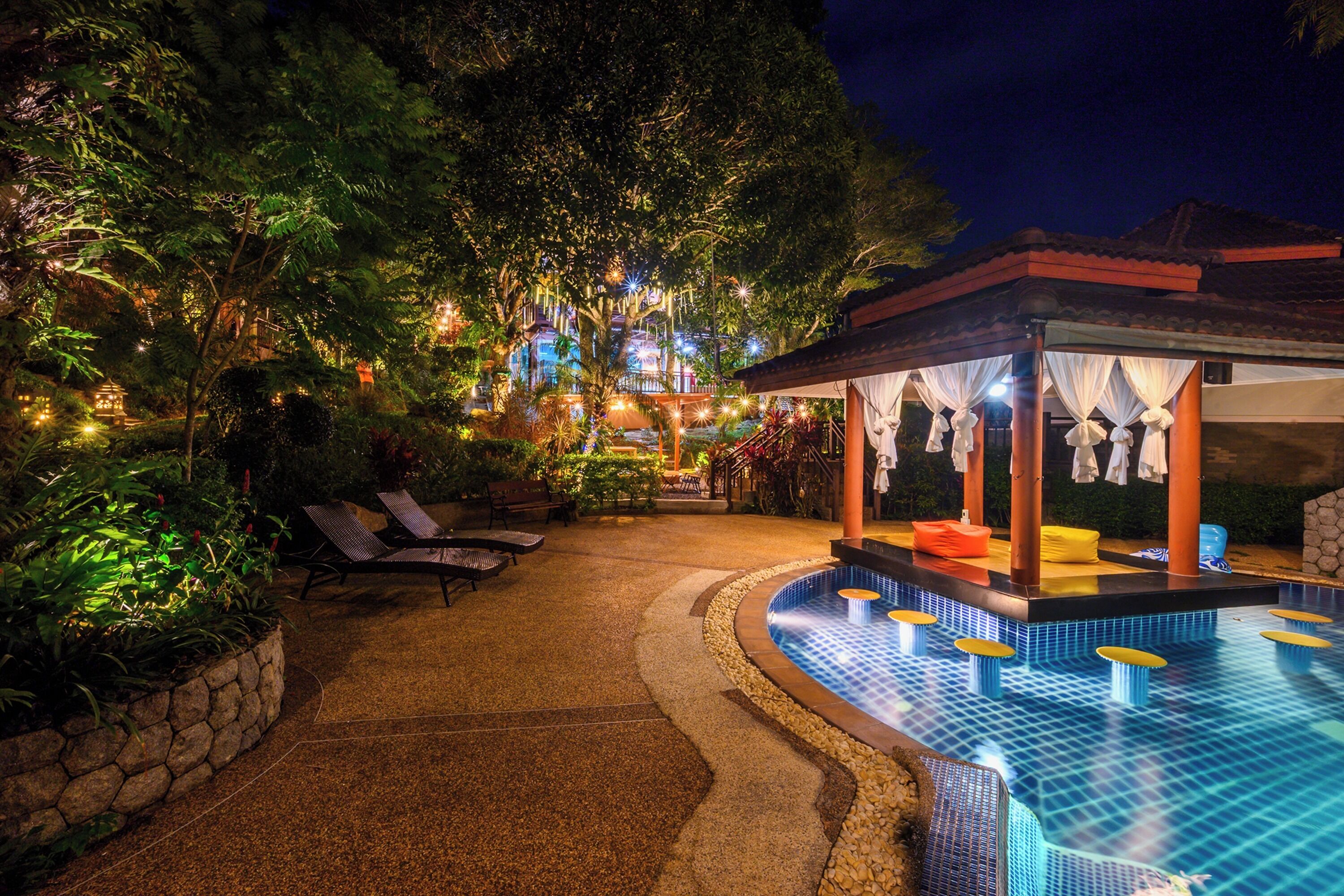 Ein Poolbereich bei Nacht mit Liegestühlen, einer überdachten Sitzgelegenheit und einem gut beleuchteten Gebäude im Hintergrund.