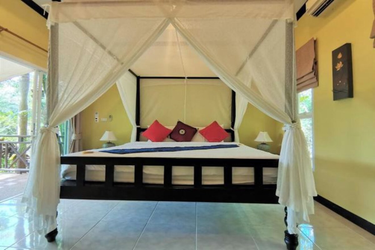 A bed with a canopy, pillows, and a bedside table in a room with tiled flooring and a yellow wall.