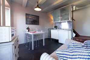 A compact living space with a kitchenette, a dining area with transparent chairs, a bed with patterned bedding, and a painting on the wall.