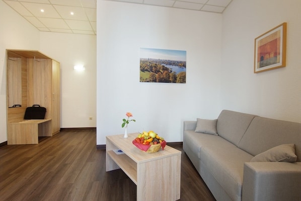 Una habitación moderna con un banco de madera, una mesa de madera con frutero y un florero, un sofá gris y un cuadro de paisaje enmarcado en la pared.