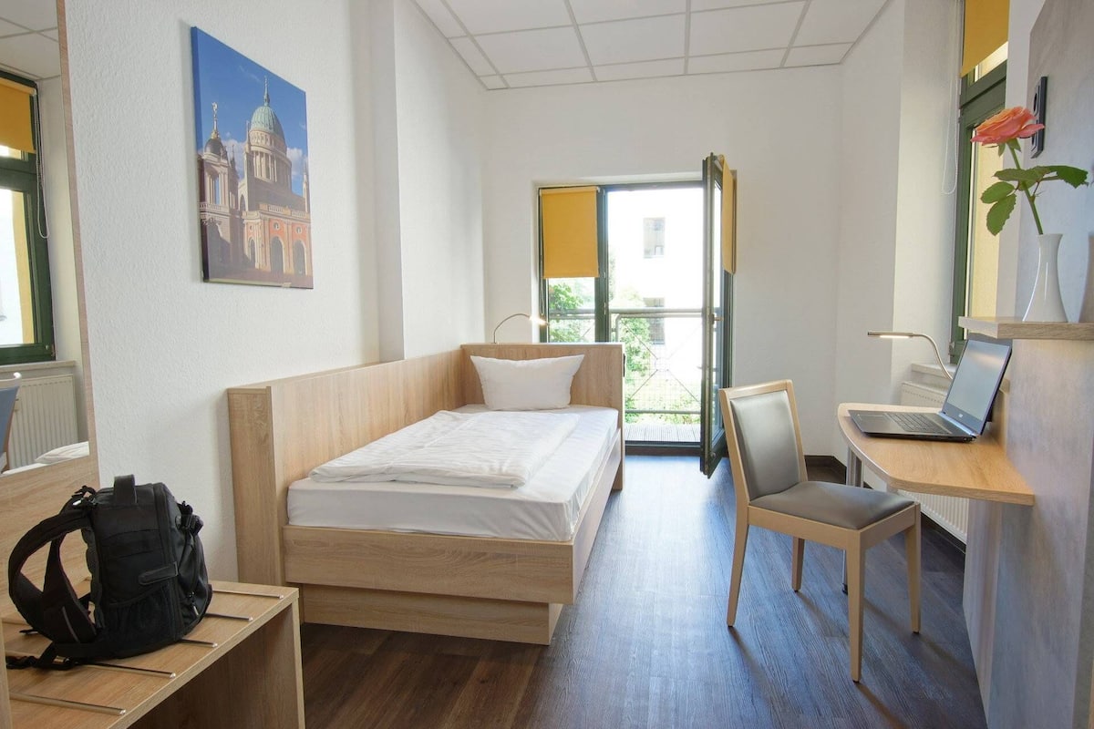 Ein Hotelzimmer mit einem Bett, einem Schreibtisch mit einem Laptop, einem Stuhl, einem Rucksack auf einem Regal und einem Bild von einem Gebäude an der Wand.