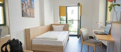A hotel room with a bed, a desk with a laptop, a chair, a backpack on a shelf, and a painting of a building on the wall.