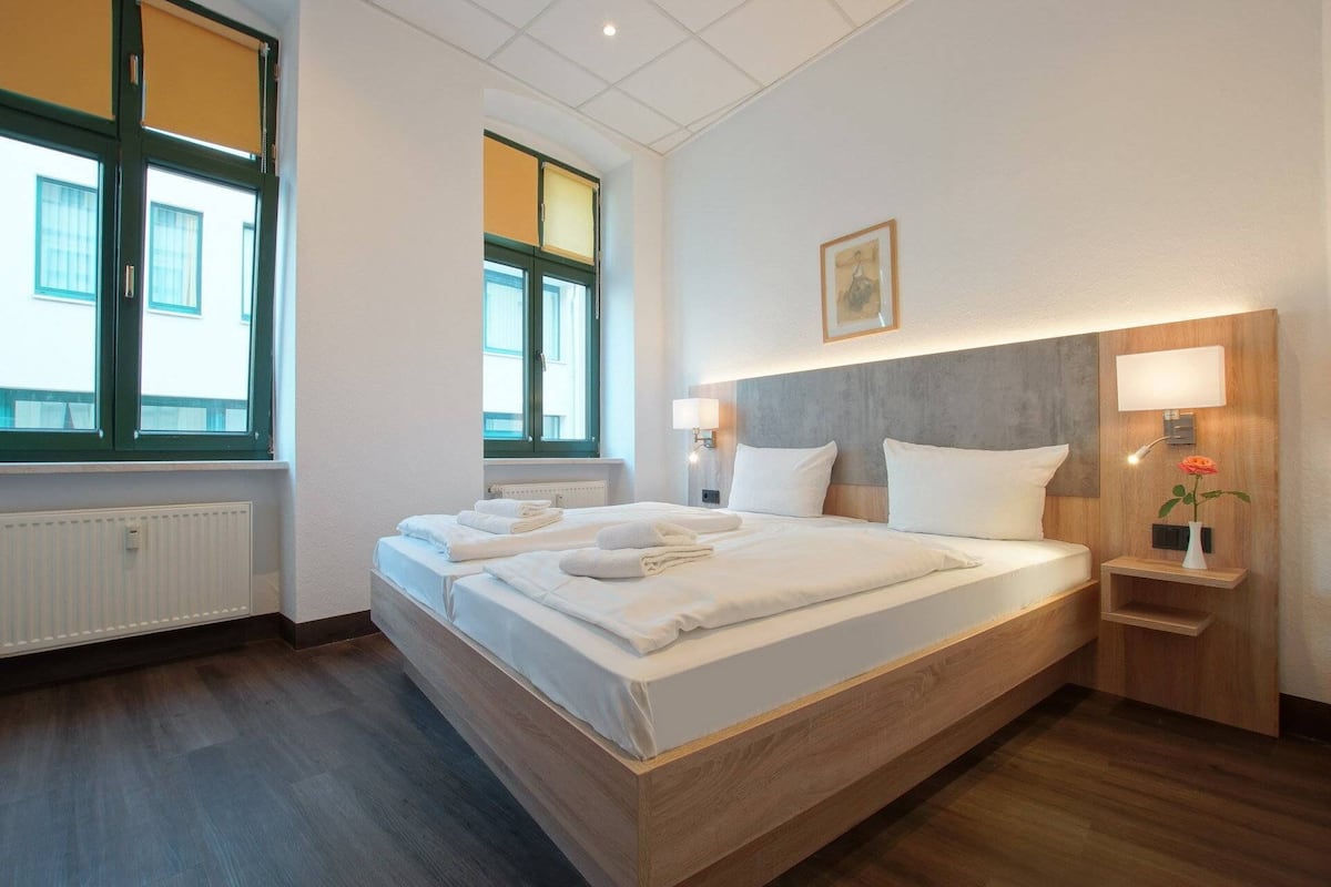 Ein Hotelzimmer mit einem großen Bett, zwei Nachttischen, einem Heizkörper und einem Fenster mit Jalousien.