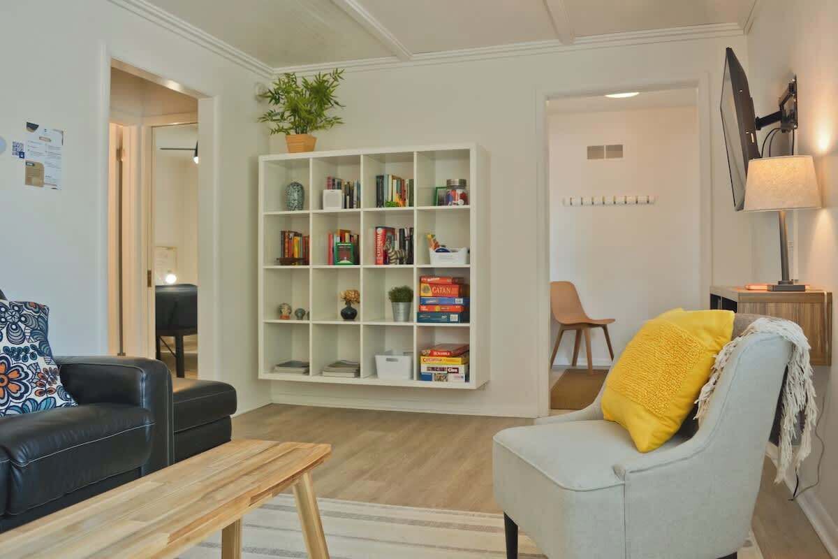 A living room with a black leather sofa, a white armchair with a yellow pillow, a wooden coffee table, and a white bookshelf filled with books and decorative items.