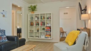Ein Wohnzimmer mit einer schwarzen Ledersofa, einem weißen Sessel mit gelbem Kissen, einem Holztisch und einer weißen Bücherwand voller Bücher und Dekoration.
