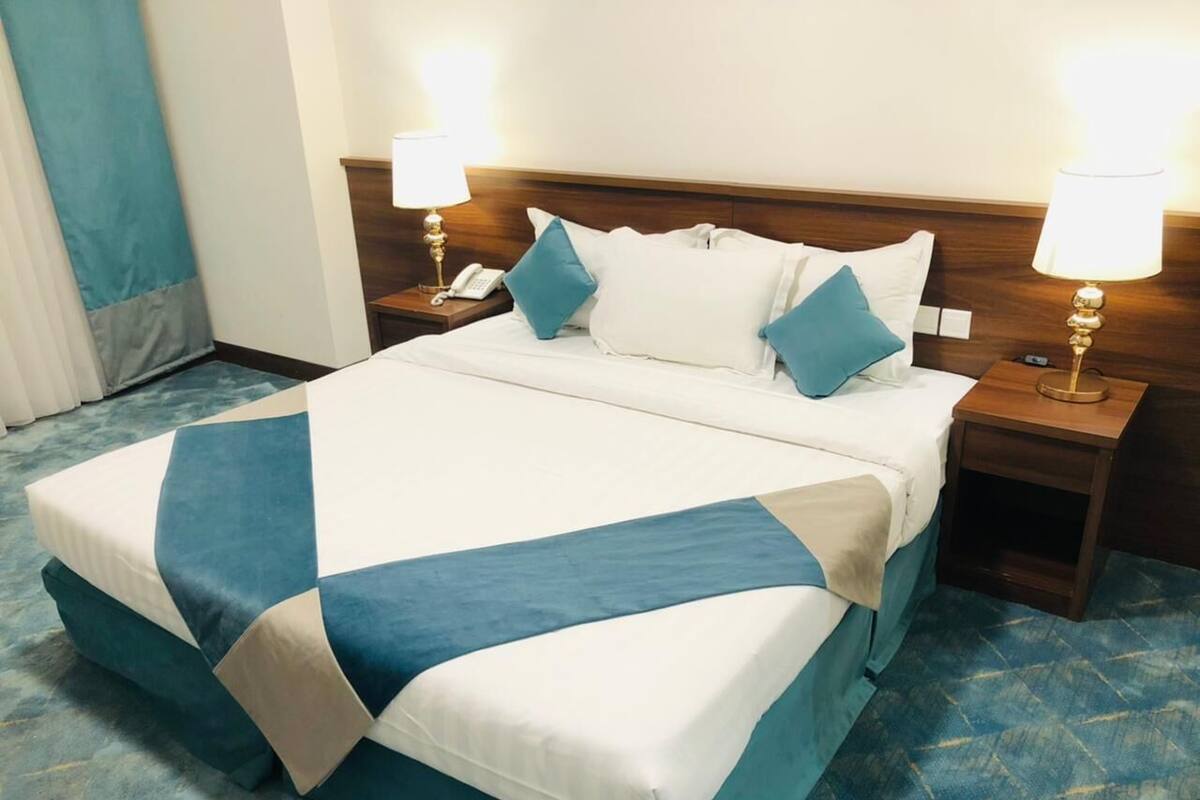 Une chambre d’hôtel avec un grand lit, deux tables de chevet avec des lampes et une moquette bleue.