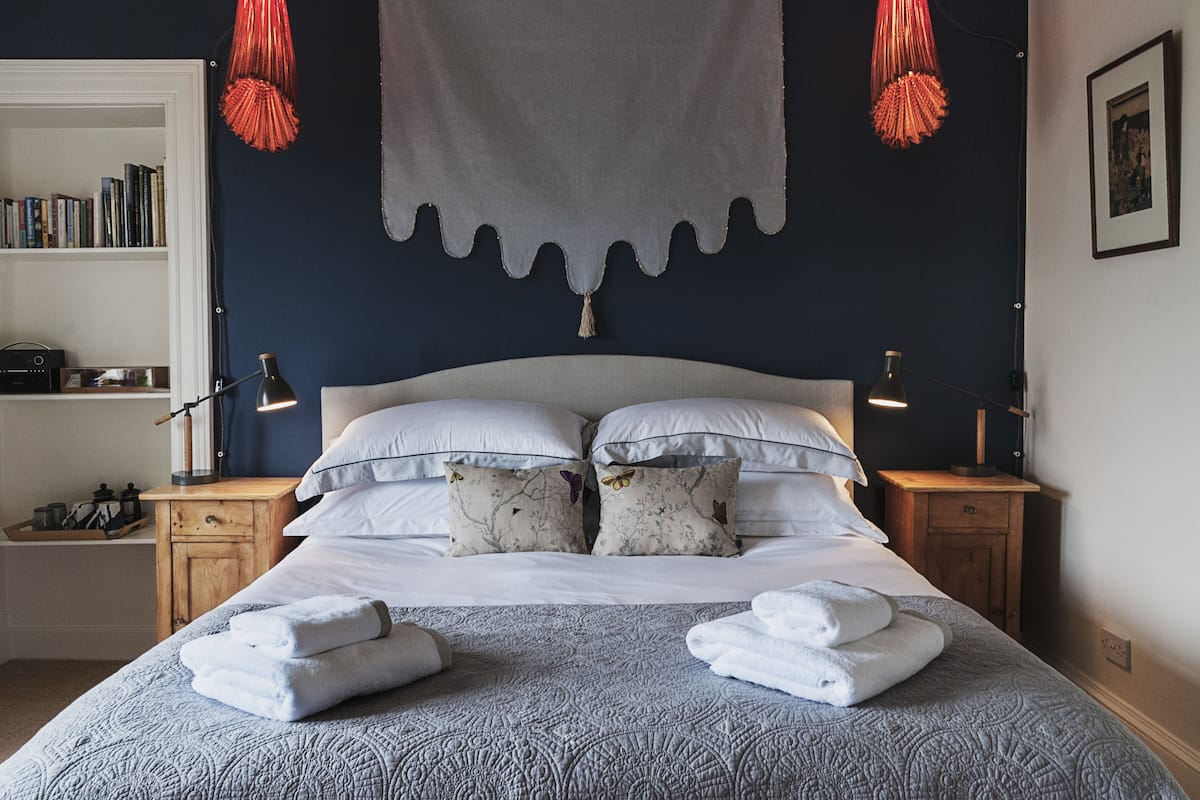 A neatly made bed with white linens and decorative pillows, a bedside table with a lamp, and a bookshelf filled with books.