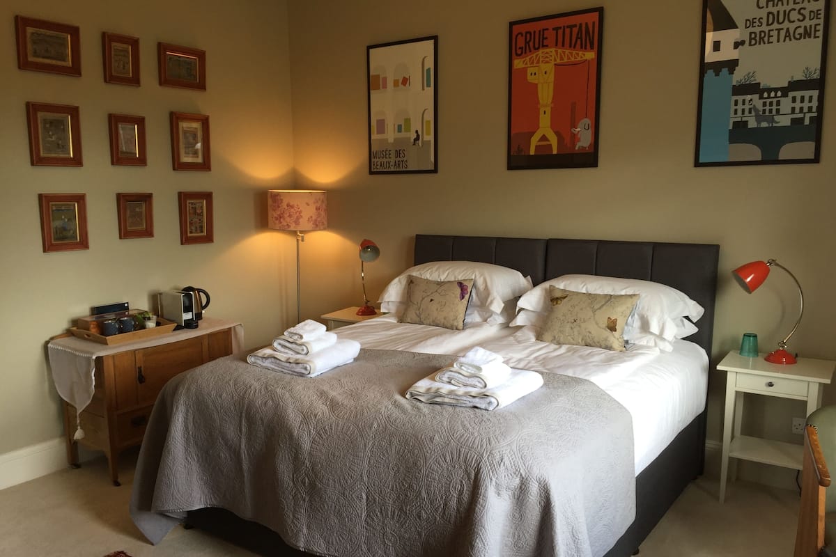 A neatly made bed with two folded towels, a bedside table with a lamp, a nightstand, and a wall with framed pictures and posters.