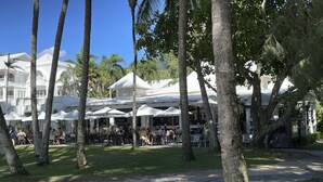Un espace extérieur aménagé pour les repas, avec des parasols blancs et des tables, entouré de palmiers et d’un bâtiment à la façade blanche.