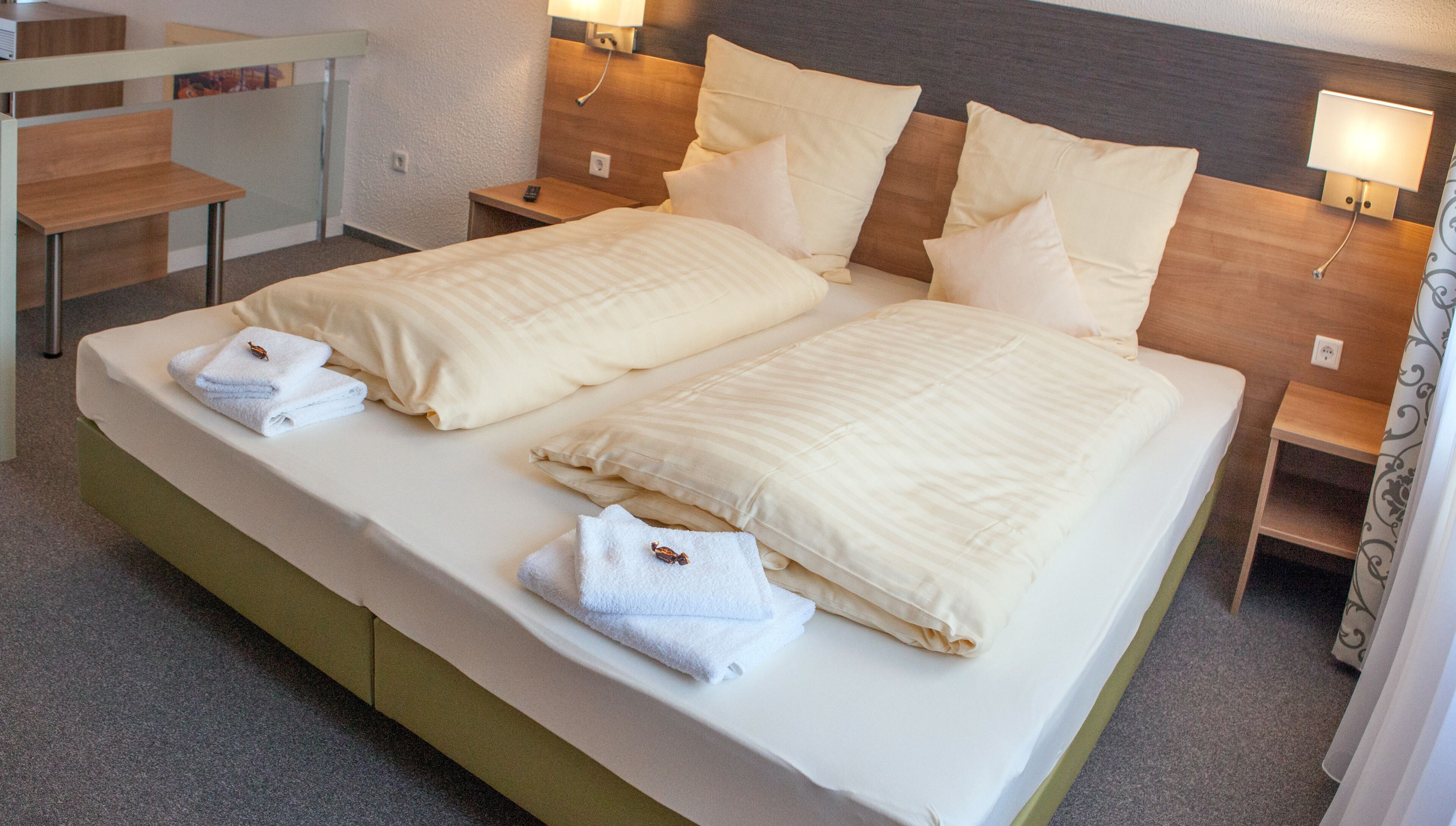 Una cama doble con sábanas y almohadas blancas, una mesita de noche con lámpara y una pequeña mesita auxiliar de madera.