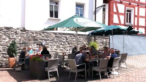 An outdoor café with wicker tables and chairs, a stone wall, and a building with a white facade and red window frames in the background. - Pension EulenNest (Laubach)