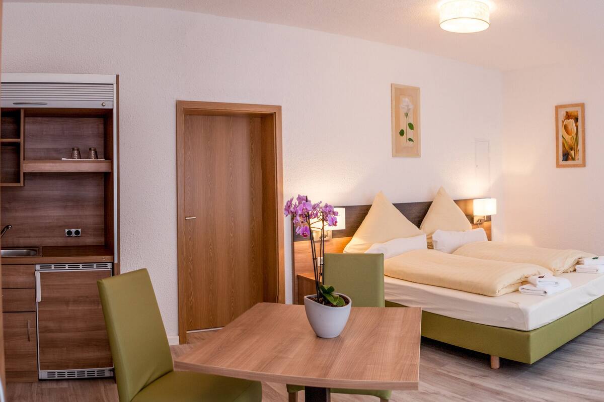 A hotel room with a bed, bedside tables, a small table with chairs, a potted plant, and a window with curtains.