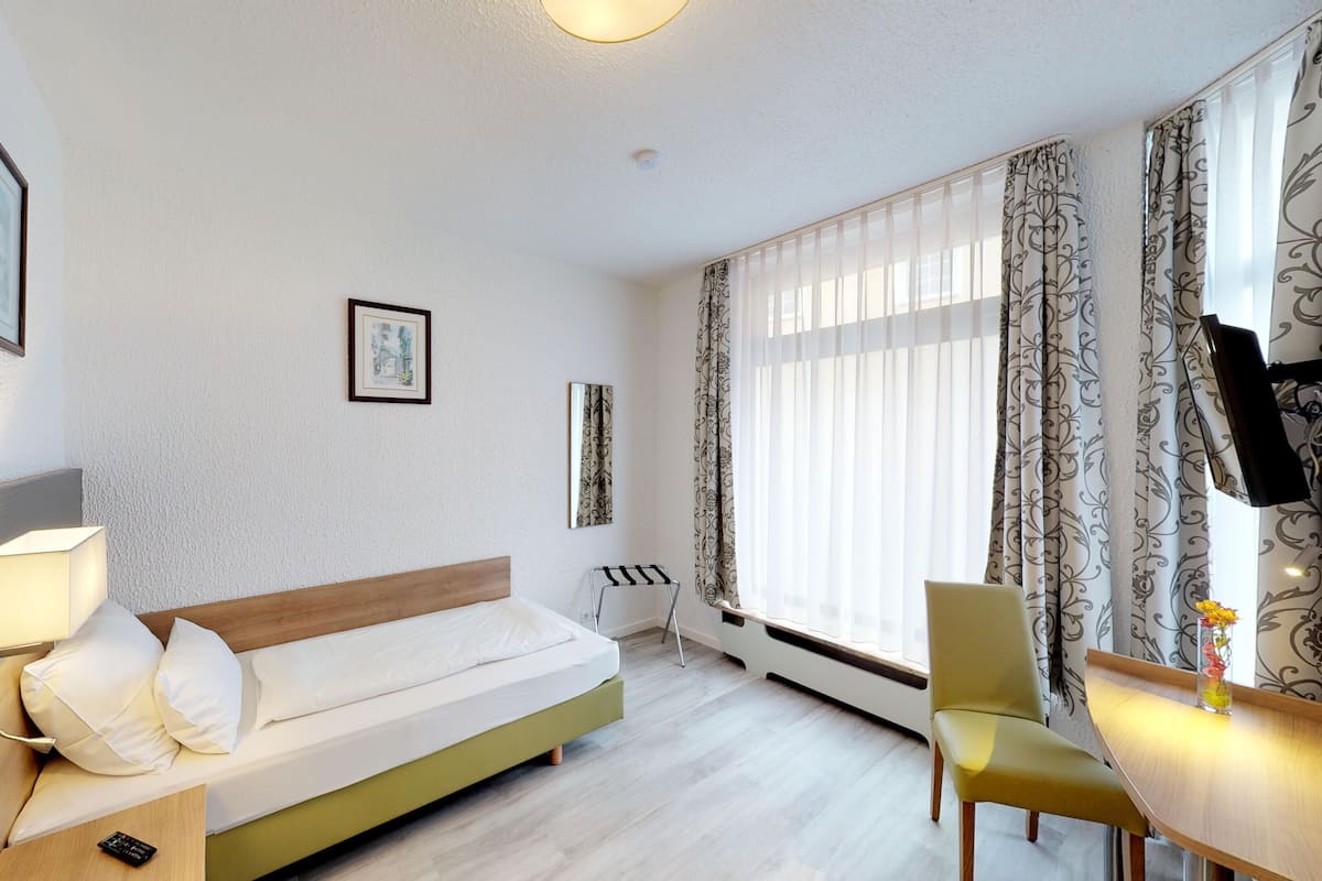 A modern hotel room with a bed, a desk, a chair, a TV, and a window with curtains.