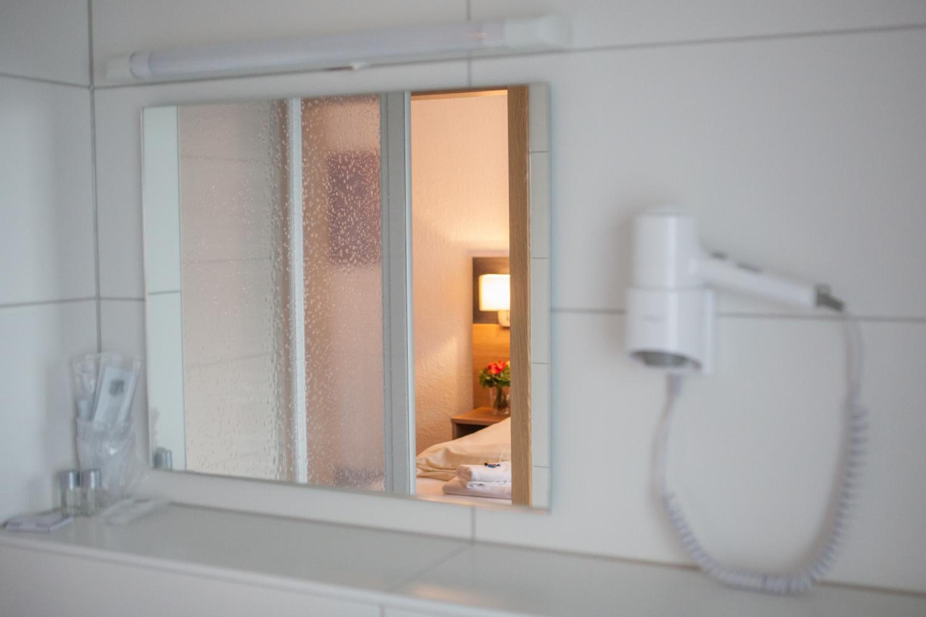 A hotel bathroom with a hairdryer mounted on the wall, a mirror with frosted glass panels, and a glimpse of a bed and a lamp in the reflection.
