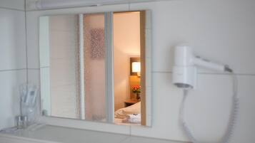 A hotel bathroom with a hairdryer mounted on the wall, a mirror with frosted glass panels, and a glimpse of a bed and a lamp in the reflection.