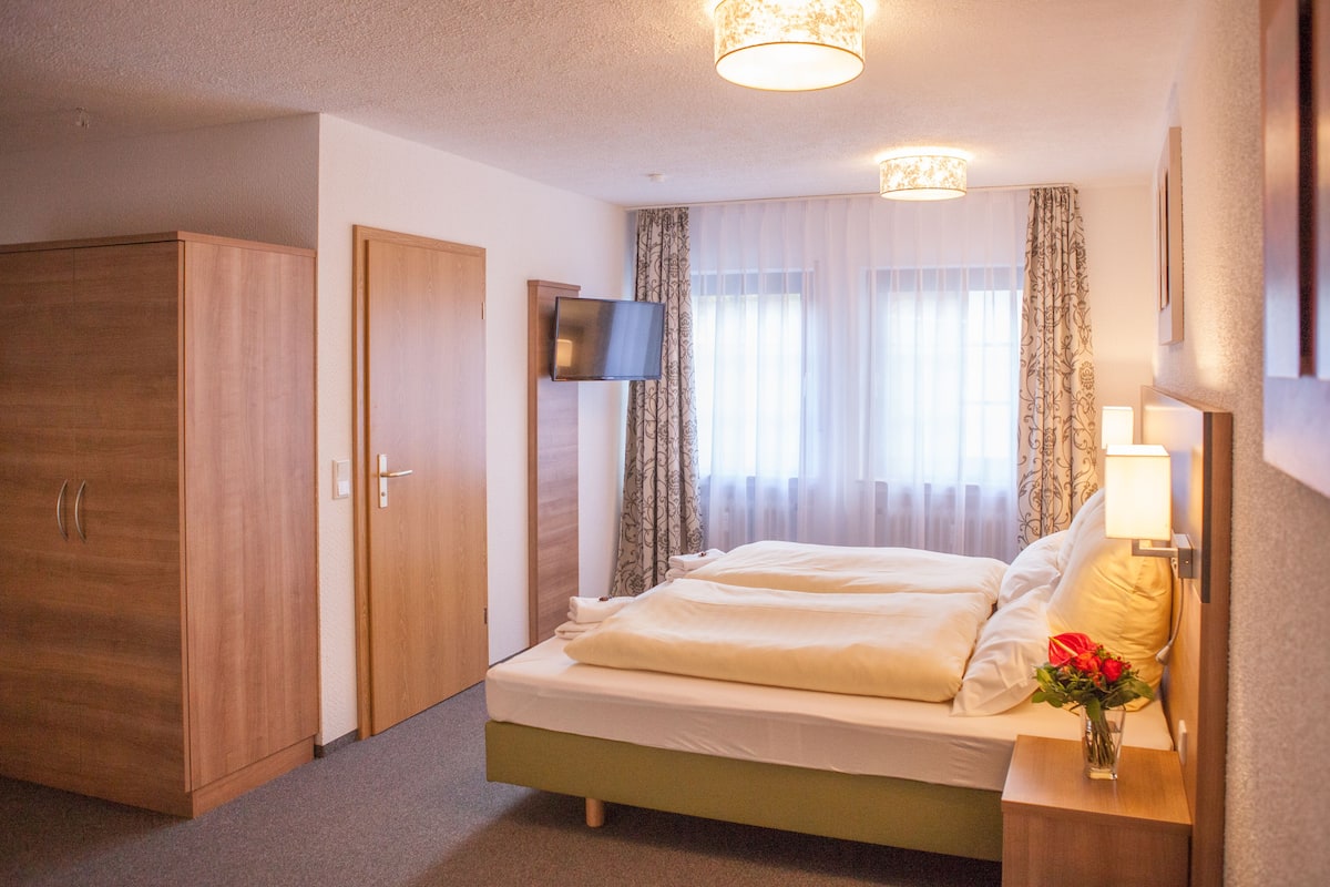 A hotel room with a bed, a wardrobe, a television, and a nightstand with a lamp and flowers.