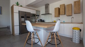 A modern kitchen with a central island, white chairs, a stainless steel refrigerator, and wooden cabinets. - Exceptional Villa with Ocean Views in Campomoro, Sleeps 6, Near the Beach (BELVEDERE-CAMPOMORO)
