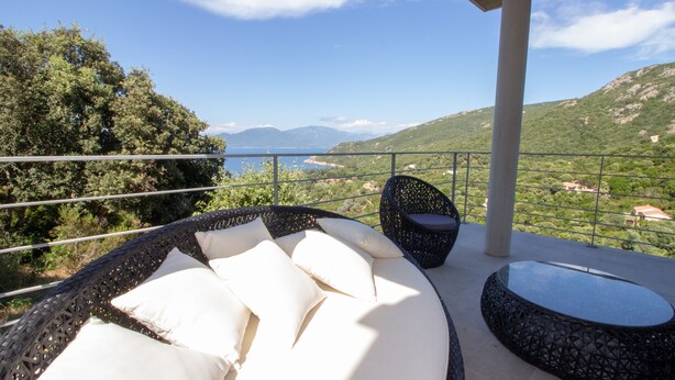 A balcony with a round black wicker sofa and a small round table, overlooking a mountainous landscape and a body of water. - Exceptional Villa with Ocean Views in Campomoro, Sleeps 6, Near the Beach (BELVEDERE-CAMPOMORO)