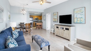 A spacious living room with a blue sofa, a patterned ottoman, and a television on a wooden cabinet. - Beachfront 2BR @ Phoenix V • Sleeps 8 • Balcony Views, Pools, Indoor Pool & Pirate Splash Pad (Orange Beach)
