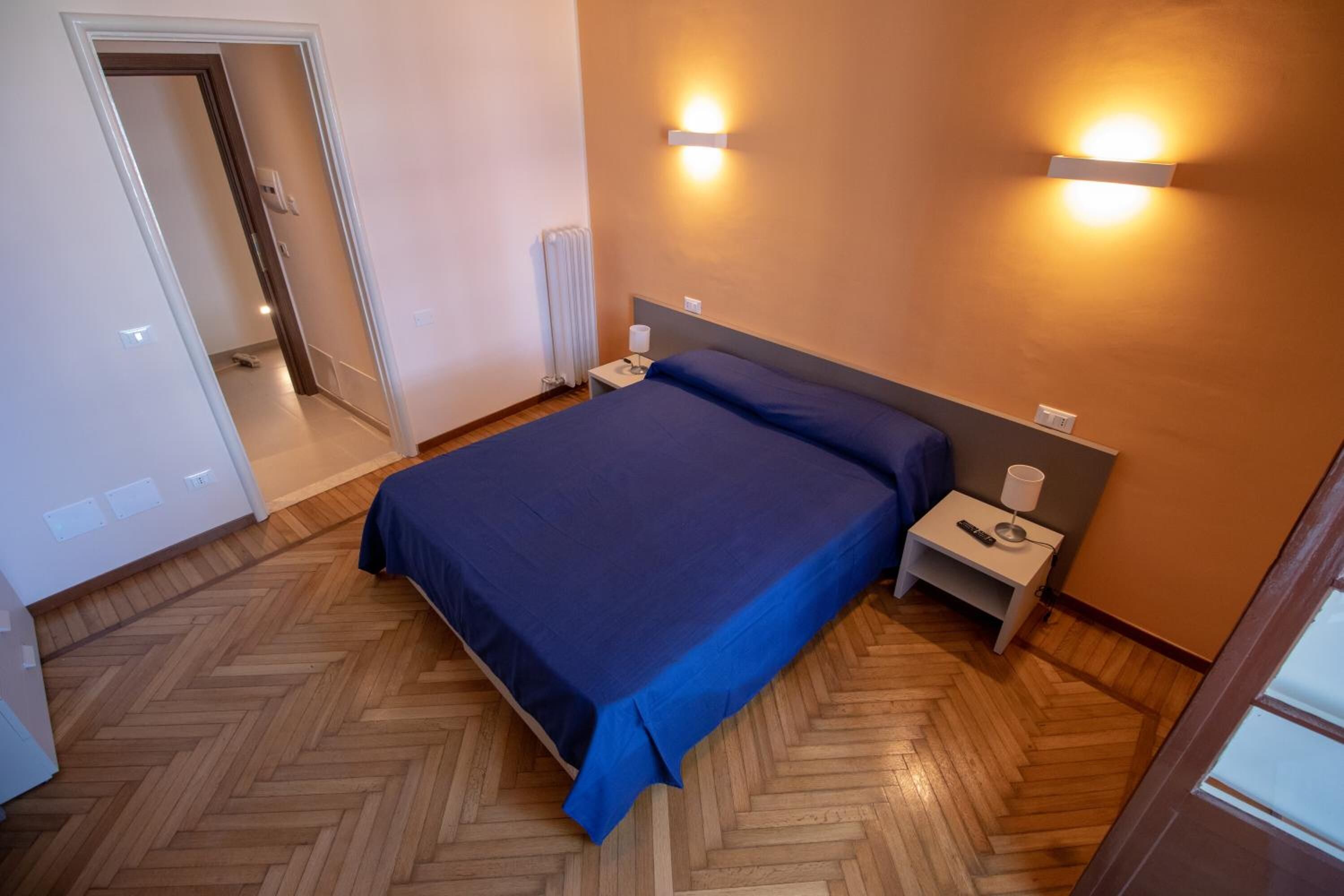 A bedroom with a bed, wooden herringbone floor, two bedside lamps, a nightstand, and a wall-mounted light fixture.