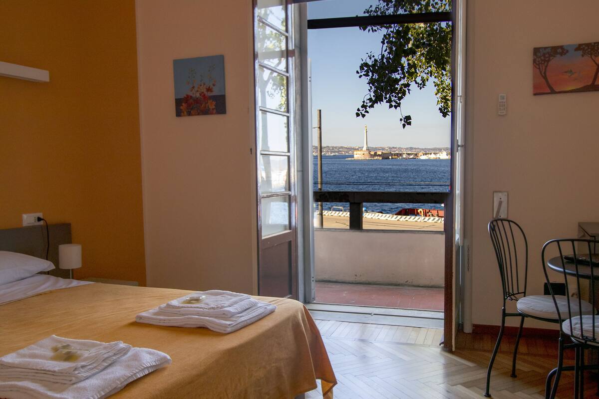 A hotel room with a bed, a chair, and a view of the sea through an open door.