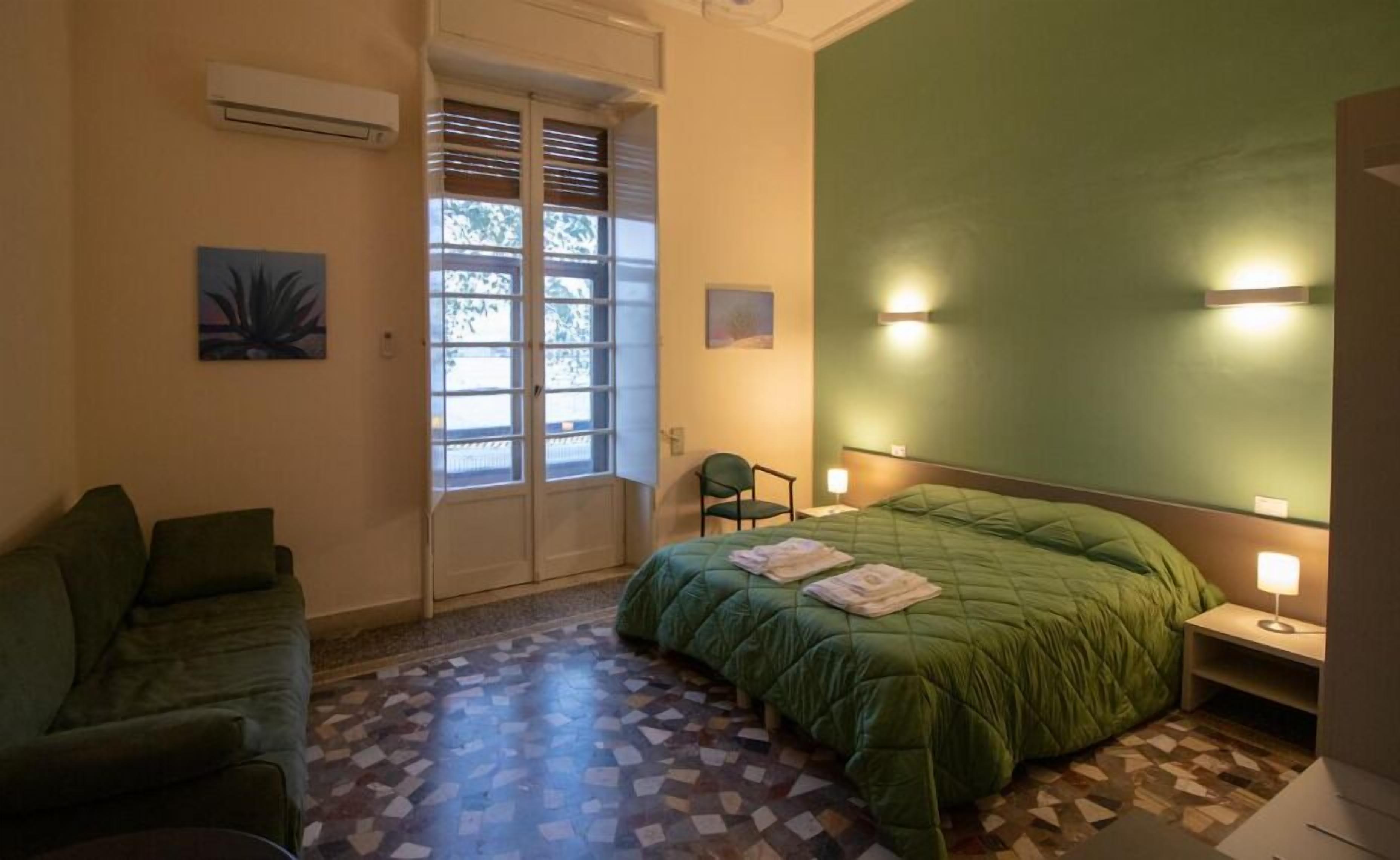 A hotel room with a green bed, a green sofa, a tiled floor, a window with blinds, and a wall-mounted air conditioner.
