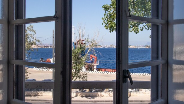 A view through a window with a clear blue sky, a body of water, and a distant shoreline with buildings and a crane. - Teatro sullo Stretto (Messina)