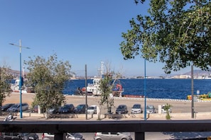 Vista costera con un barco en el puerto, coches aparcados y un cielo azul despejado.