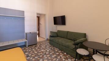 Um quarto pequeno e mobiliado com sofá, uma mesinha com cadeiras, um banco e uma televisão na parede.