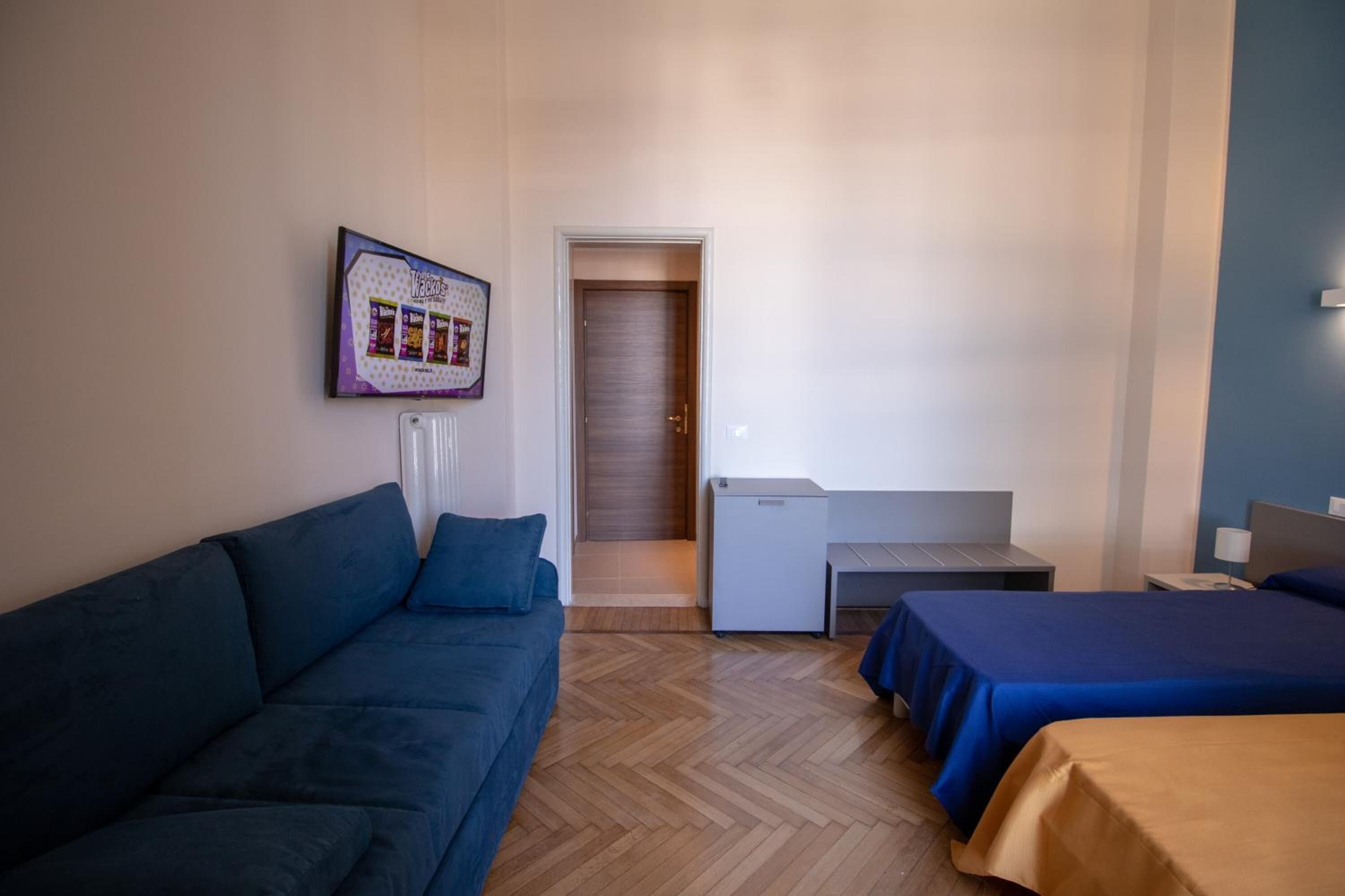 Ein Hotelzimmer mit blauer Couch, zwei Betten, einer kleinen Küchenzeile und einer Tür zu einem Flur.