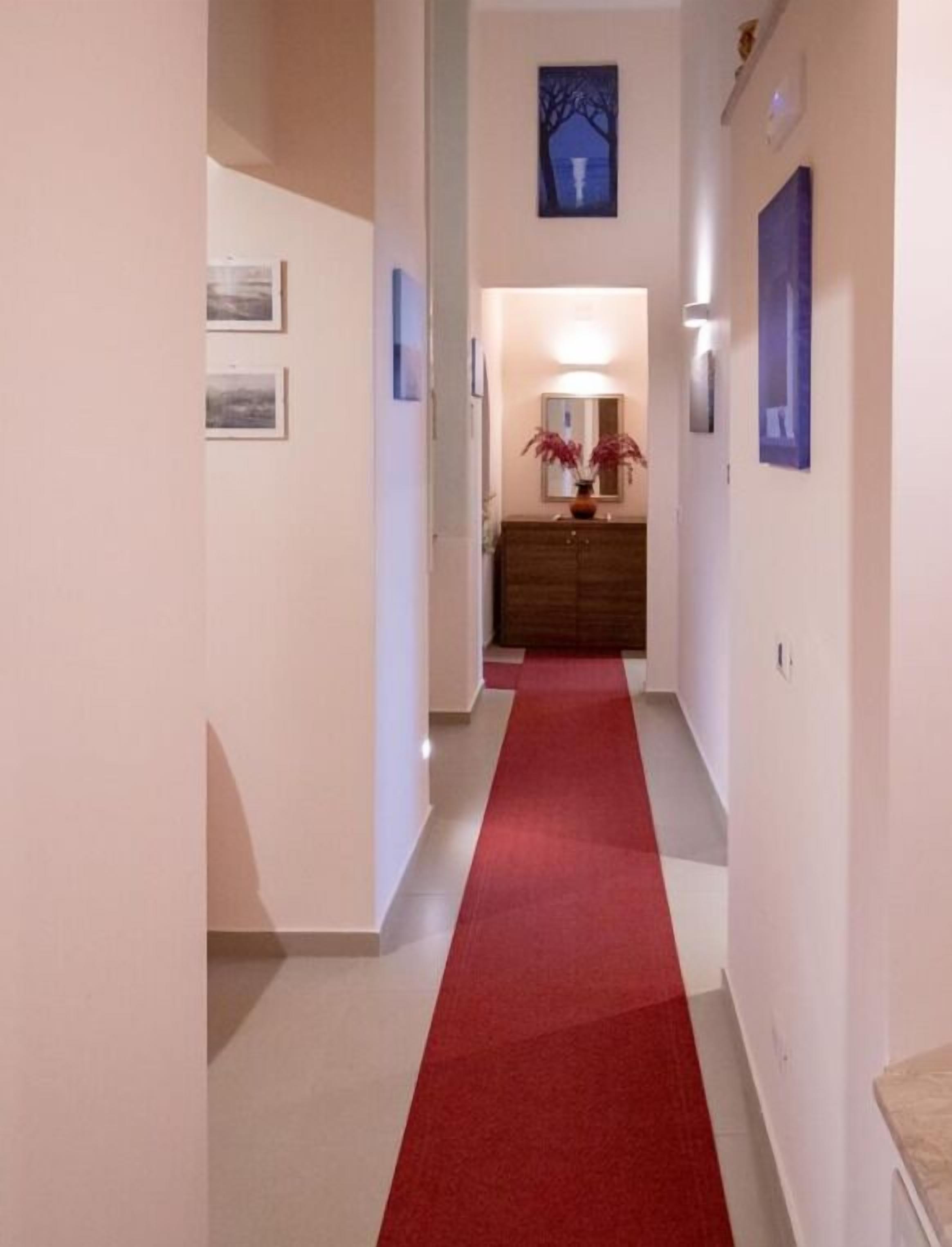 Un pasillo con alfombra roja que lleva a una habitación con un aparador de madera y un florero con flores.