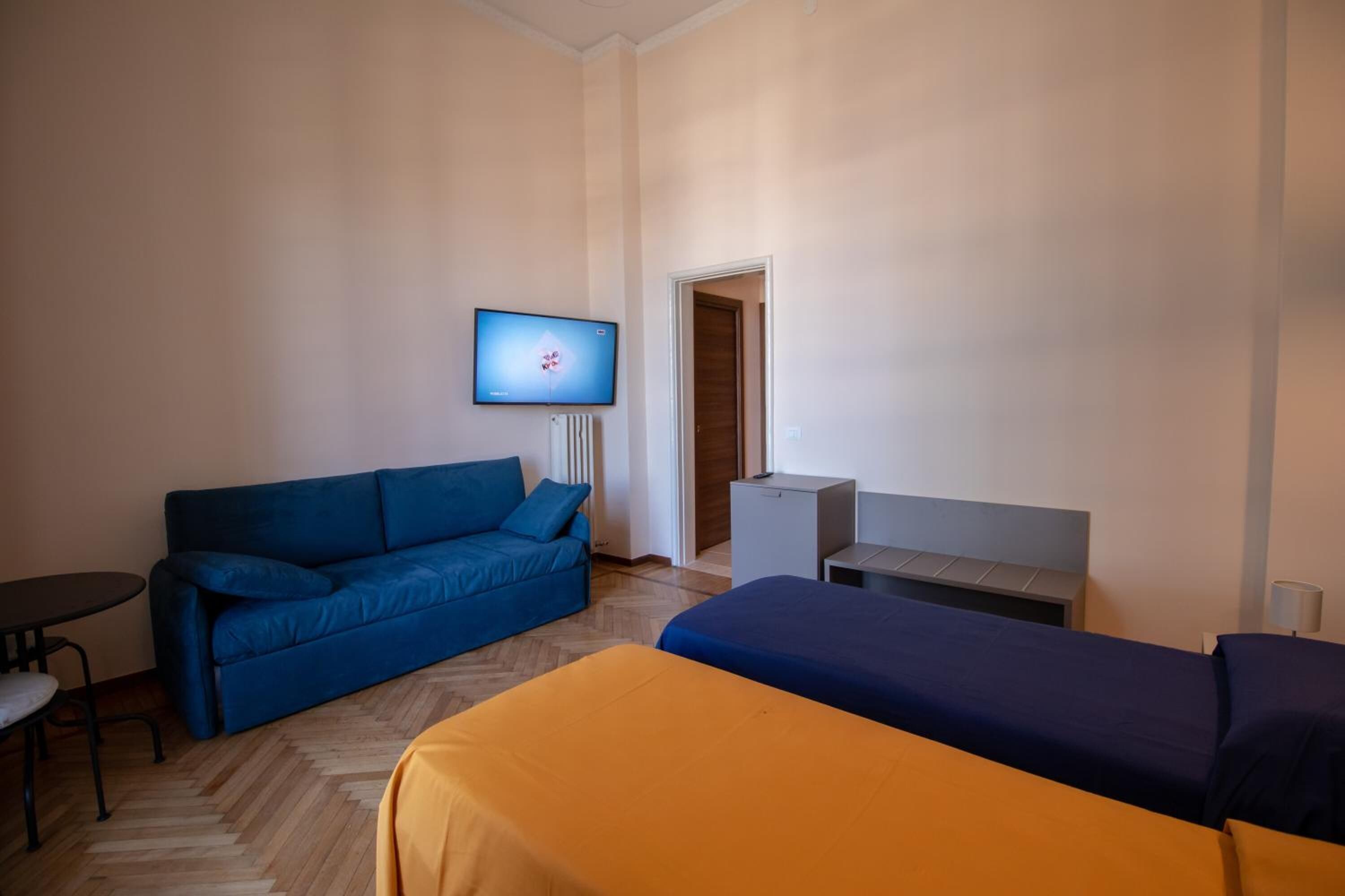 Habitación de hotel con un sofá azul, una cama naranja, una mesita pequeña, un televisor y una puerta que da a otra habitación.