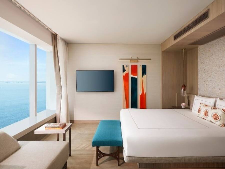 Habitación de hotel con una cama grande, una mesita de noche, un banco, un televisor montado en la pared y una ventana con vista al mar.