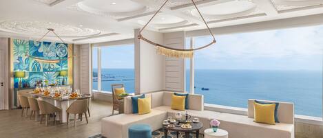 Amplia habitación con un ventanal grande con vista al mar, un comedor con mesa dispuesta para cuatro personas, y una sala de estar con sofás y una mesa de centro.