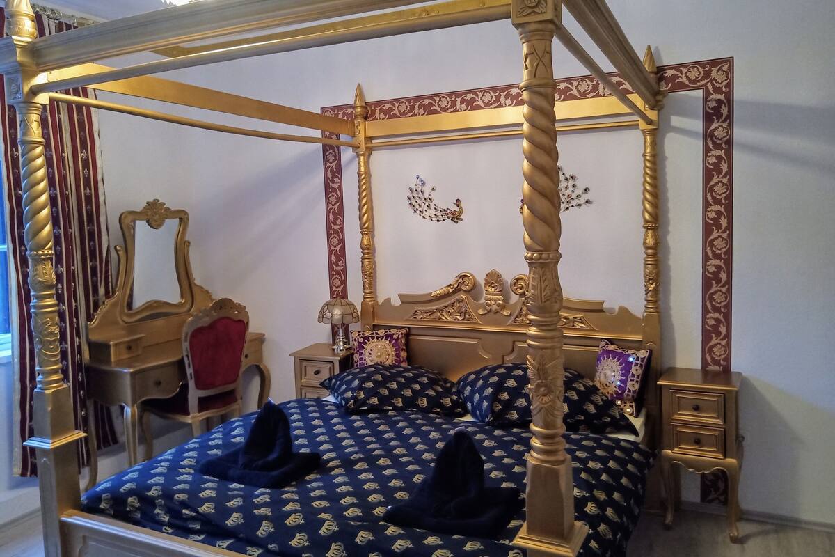 A four-poster canopy bed with a gold frame, a mirror, a red chair, and a bedside table with a lamp.