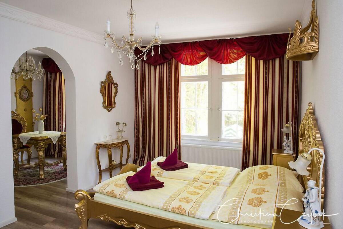 A luxurious bedroom with a large bed, ornate gold furniture, and a chandelier.