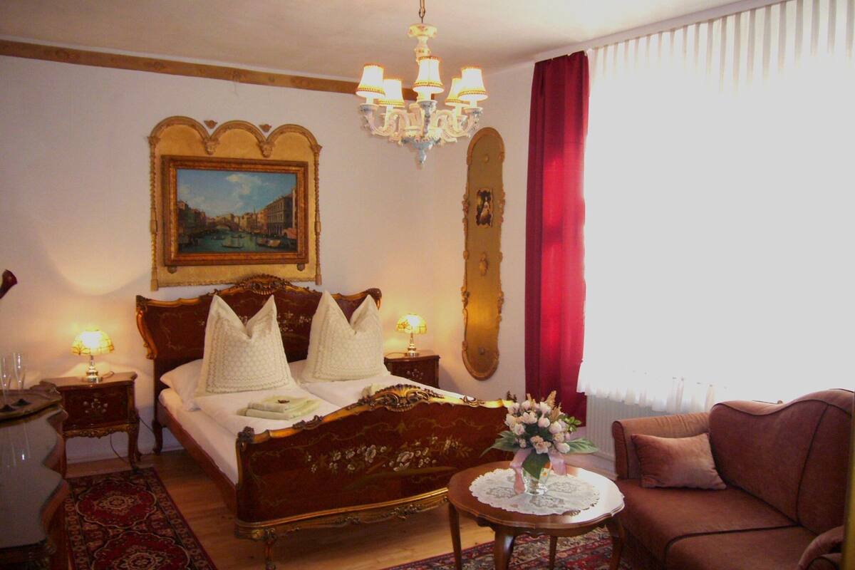 A hotel room with a large bed, a chandelier, a painting, a mirror, a red curtain, a small table with a vase, and a sofa.