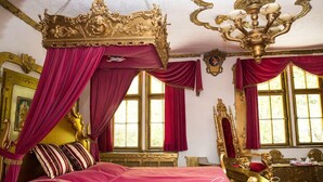 Un dormitorio lujoso con cama con dosel, detalles dorados ornamentales y cortinas rojas.