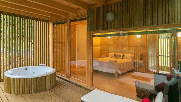 A wooden deck with a hot tub, a glass-enclosed bedroom with a bed, and a wicker basket. - Enoturismo María Manuela (Boqueixon)