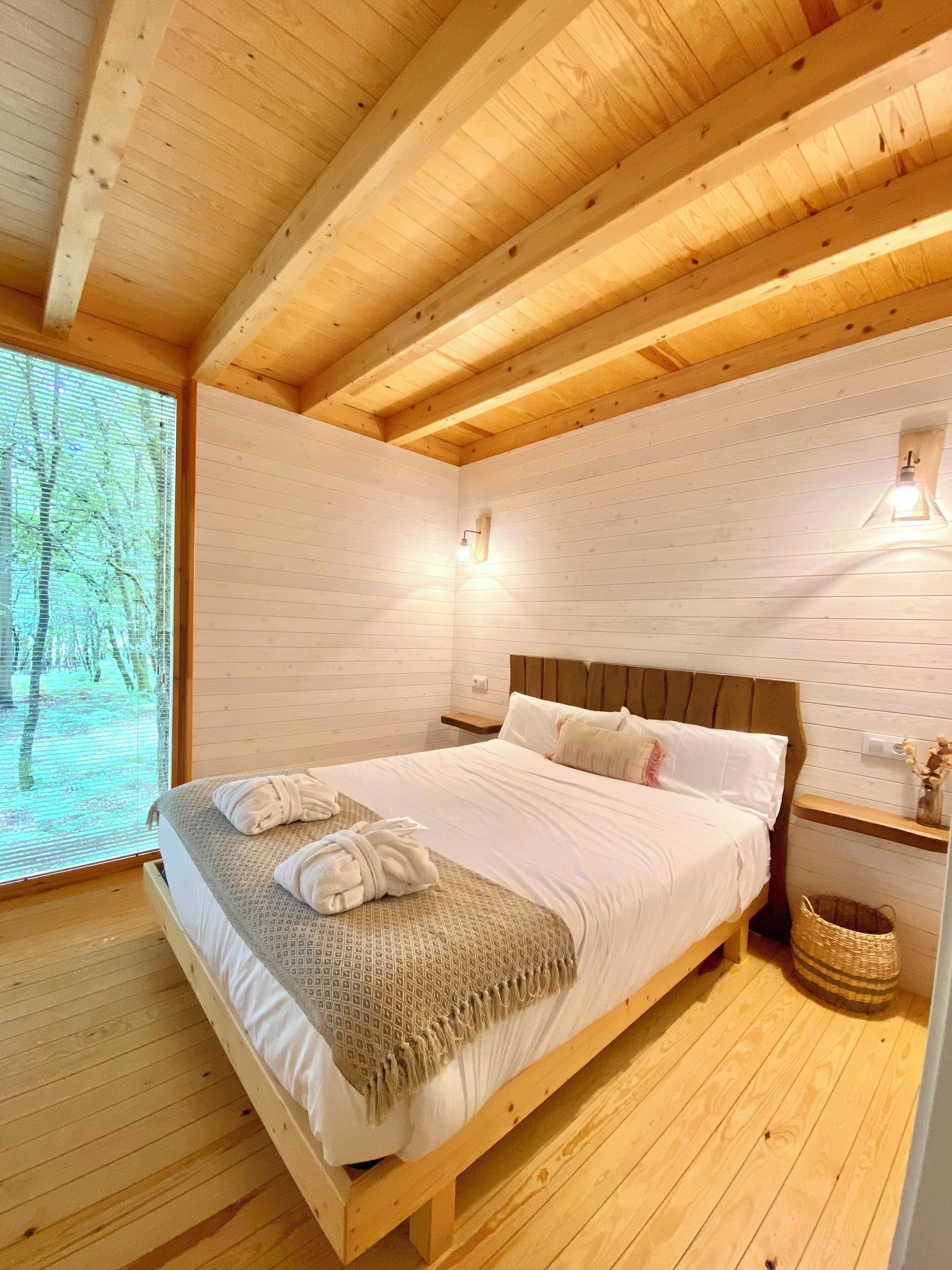 Une chambre dans une cabane en bois, avec un lit, deux tables de chevet, une fenêtre avec des stores et un luminaire fixé au mur.