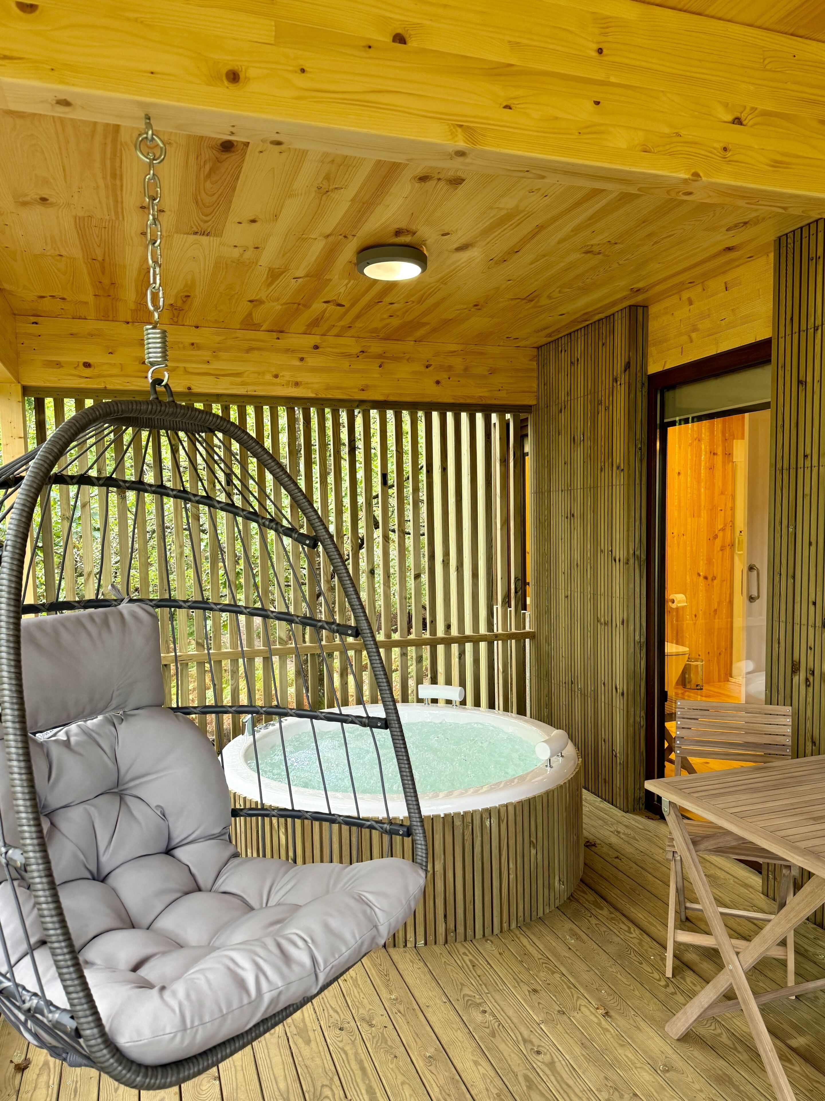A wooden deck with a hanging chair, a hot tub, and a table.