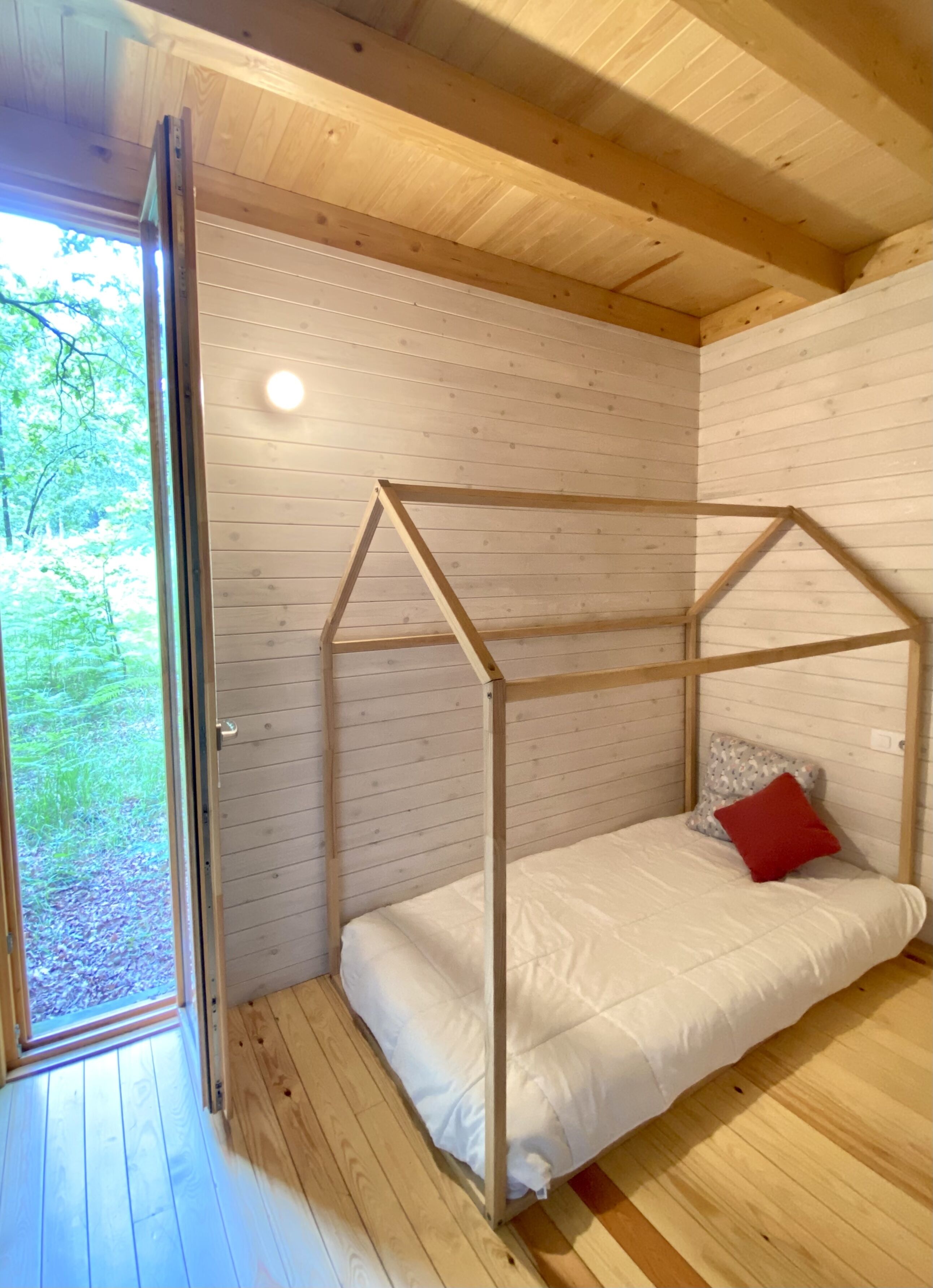 A wooden cabin room with a bed under a wooden frame, a window, and a door.